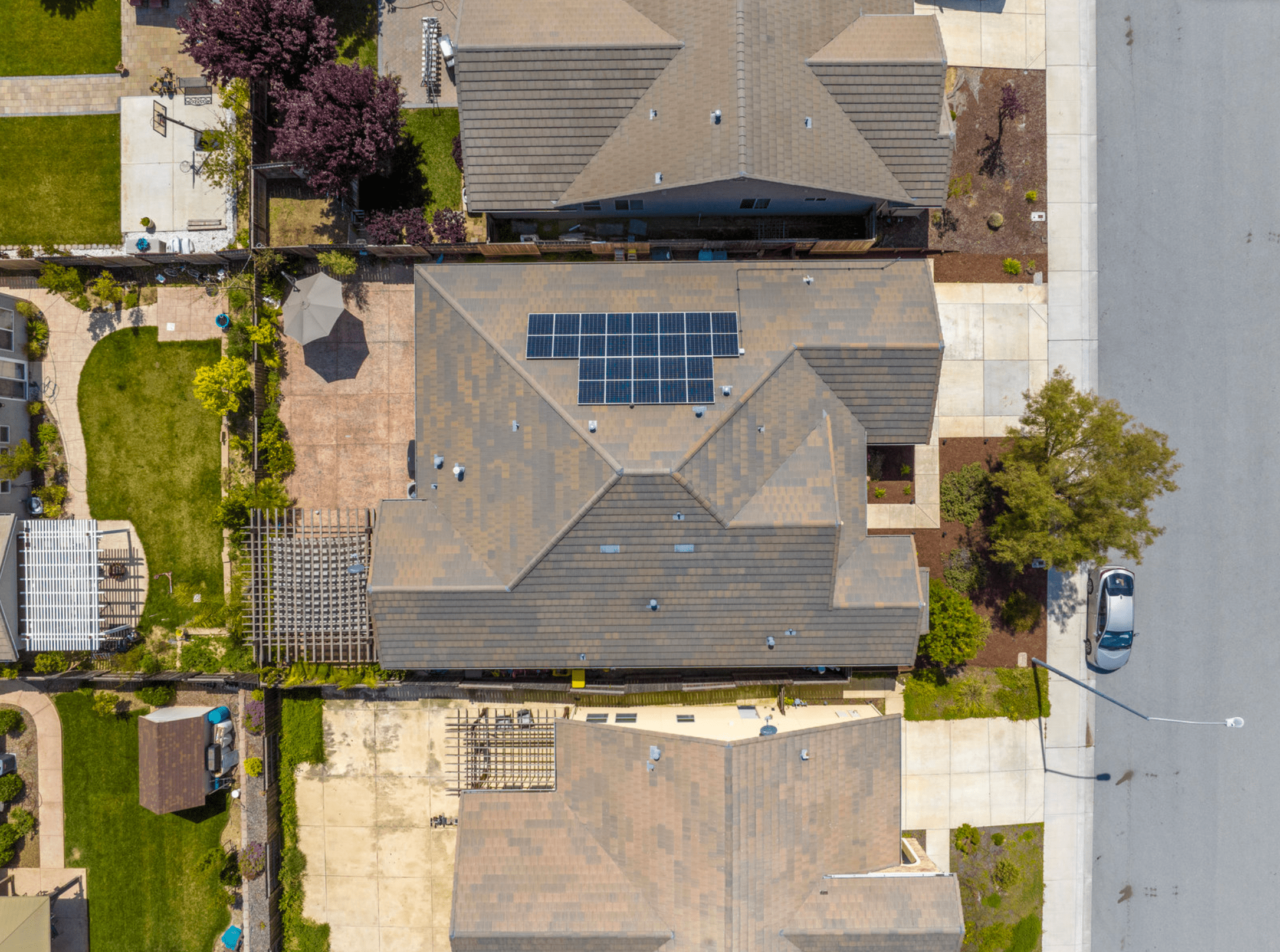 Vue aérienne d'une maison résidentielle équipée de panneaux solaires sur son toit. L'image montre une installation photovoltaïque bien intégrée sur une toiture en pente. On peut également apercevoir le jardin soigné et les maisons voisines, illustrant un quartier résidentiel typique adopting l'énergie solaire.